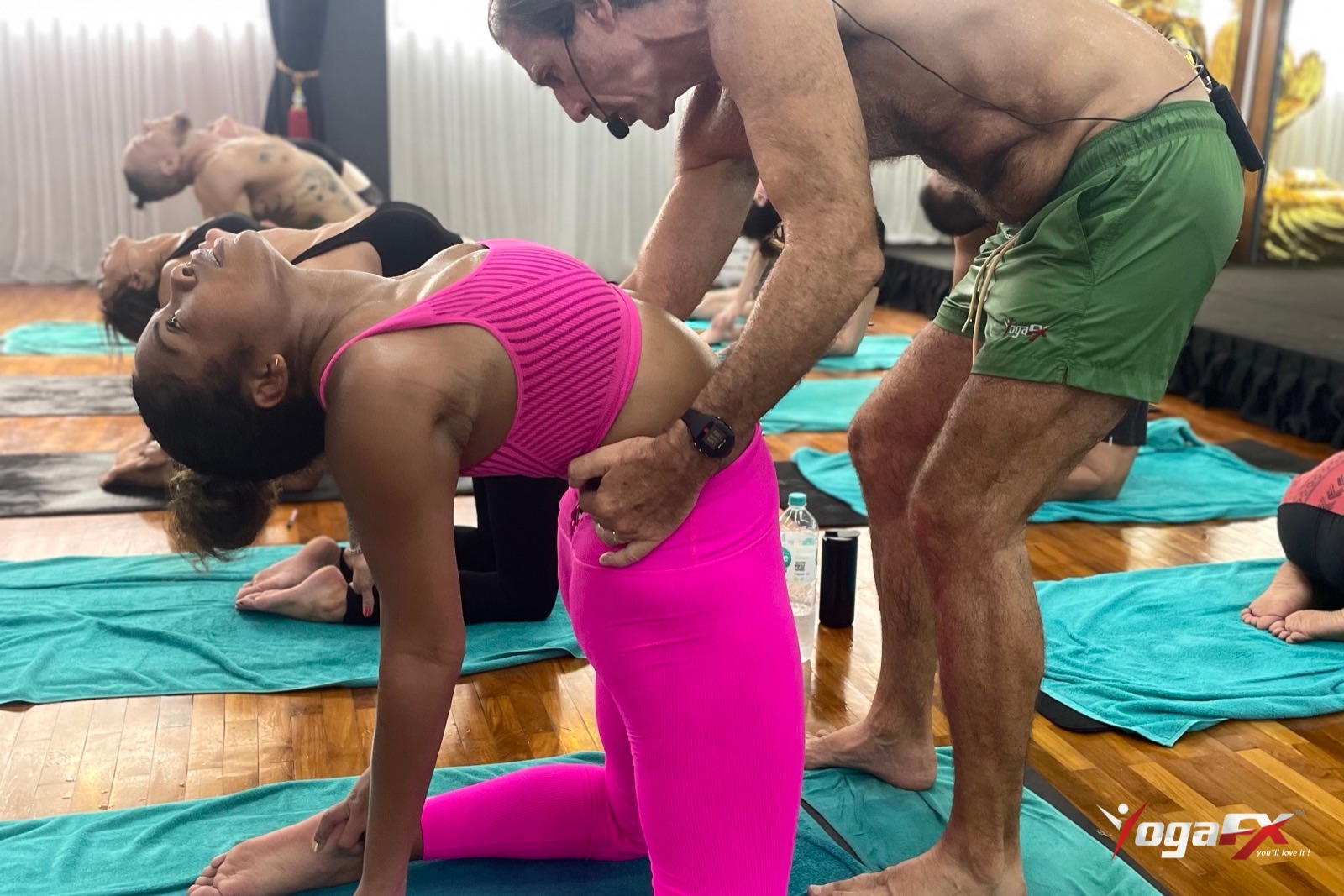 Teacher adjusting a smiling student in a kneeling backbend, the kind of hands-on guidance taught in hot yoga teacher training