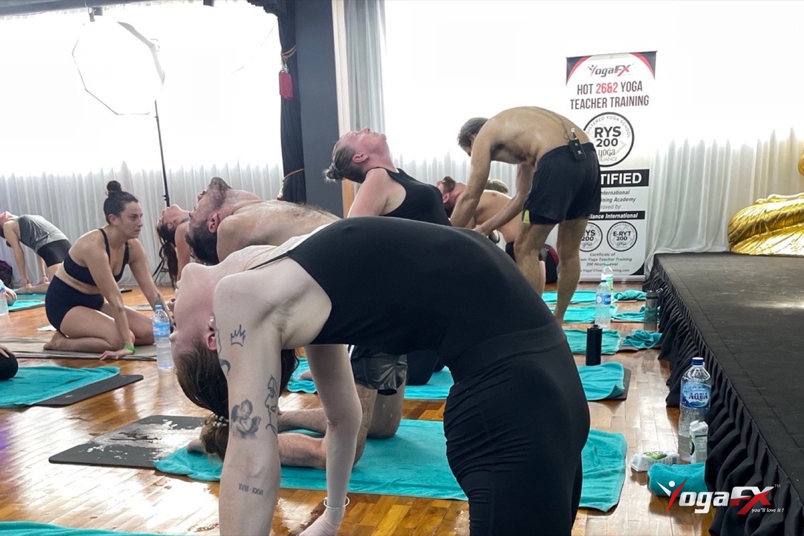 Students in Camel pose while Mr. Ian Terry adjusts a trainee, a backbend that works the back and hip muscles in hot yoga